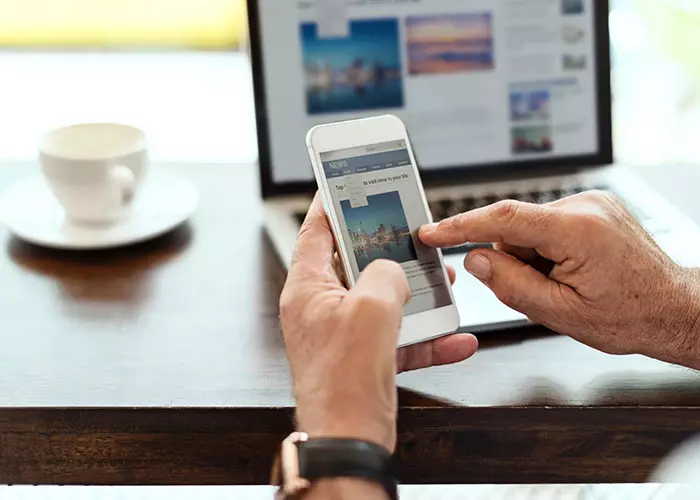 Person, die dieselbe Website auf dem Smartphone in der Hand und auf dem Laptop im Hintergrund betrachtet